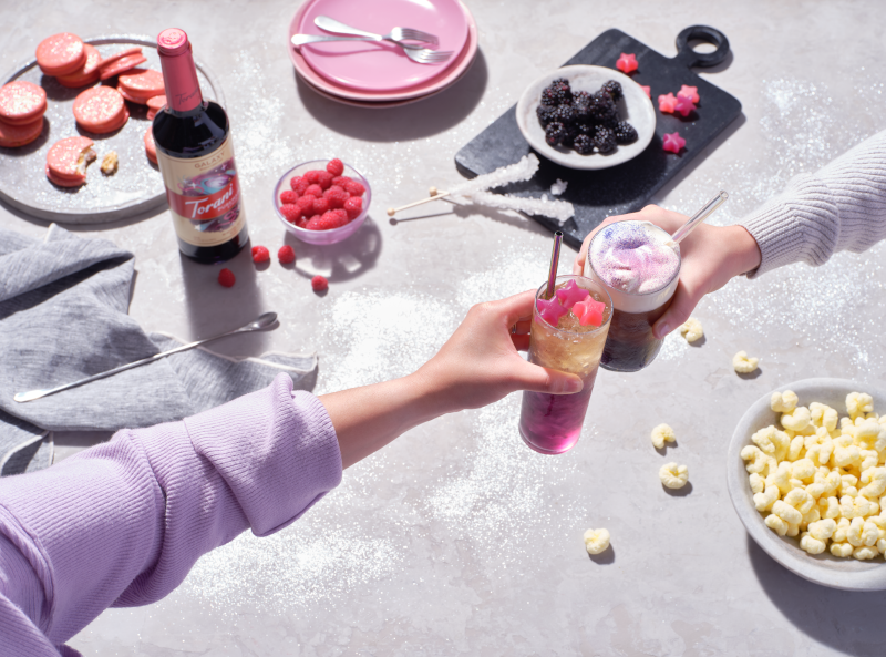 People cheersing drinks with snacks and a bottle of Torani flavored syrup in the background